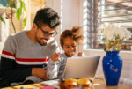 A father sits with his daughter at a desk with a laptop in front of them. She smiles as he helps her with her online schooling.